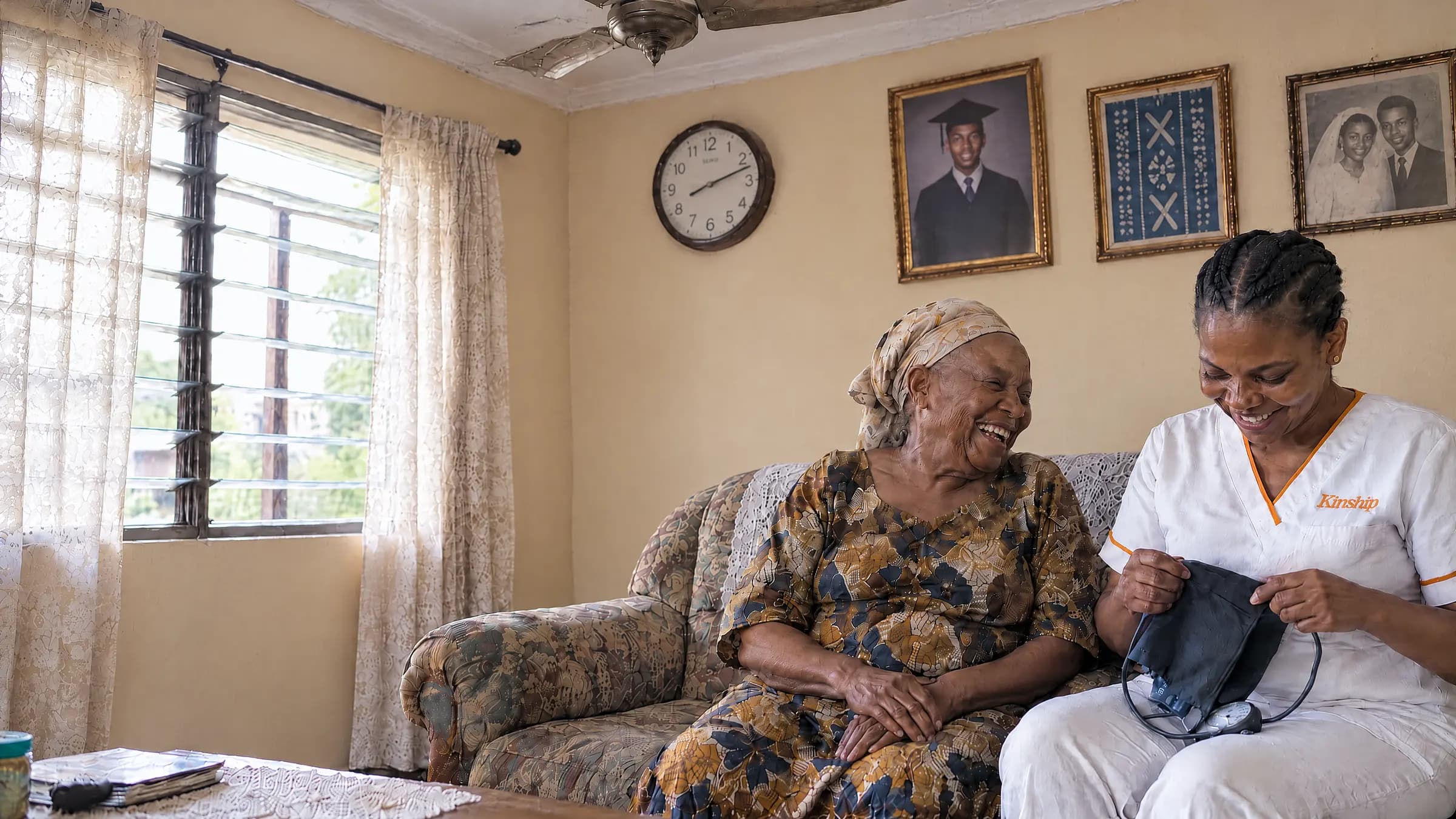 An older woman laughing on her sofa at home in Lagos as a Kinship nurse beside her unpacks a blood pressure cuff, in the morning light