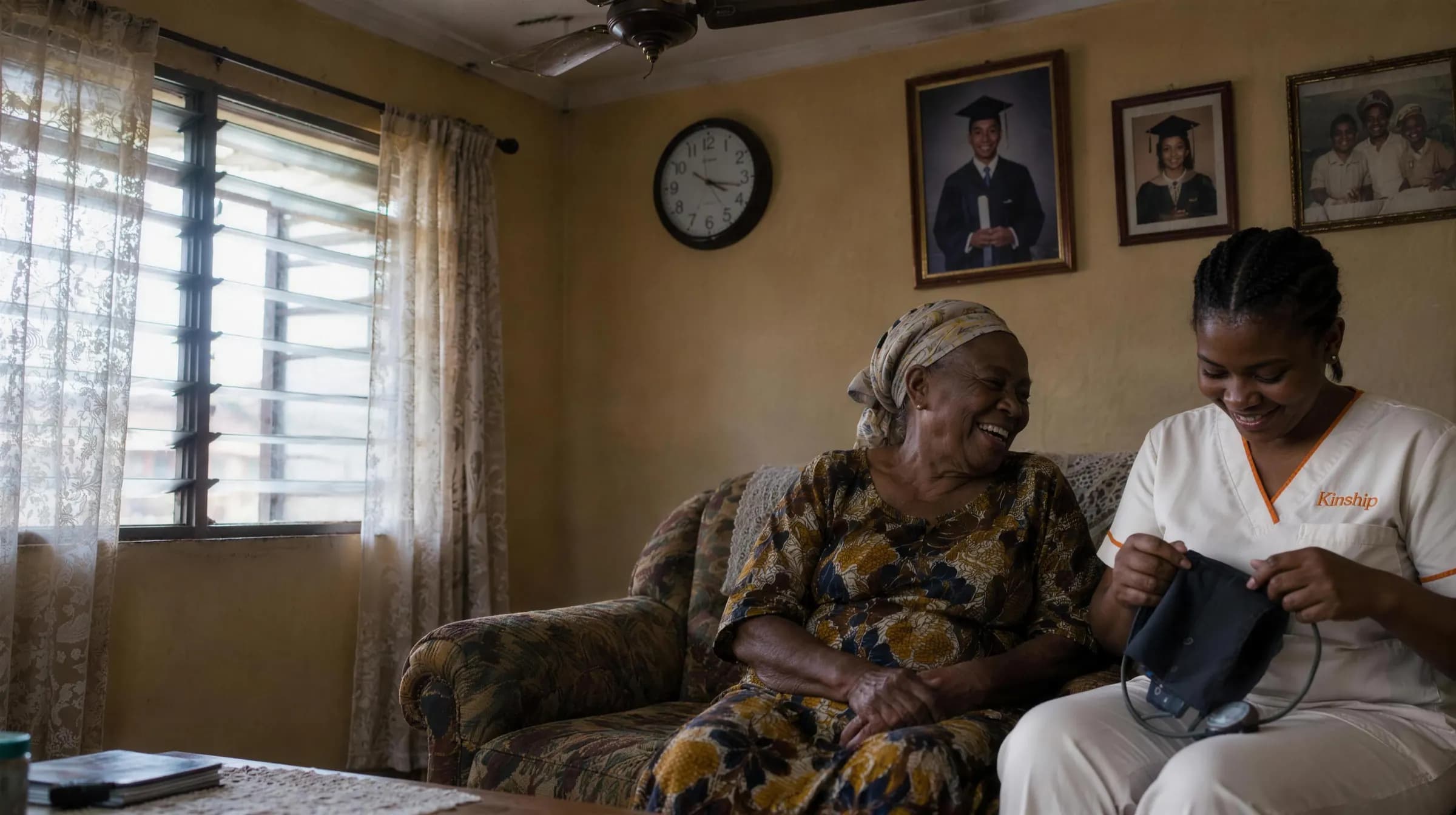 An older woman laughing on her sofa at home in Lagos as a Kinship nurse beside her unpacks a blood pressure cuff
